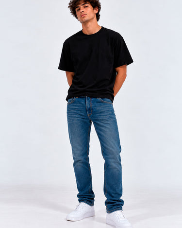 Pantalón vaquero azul claro en hombre, camiseta negra, fondo blanco, zapatos blancos.