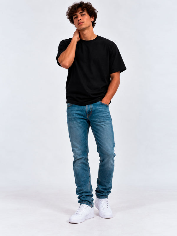 Pantalón vaquero azul claro en un hombre con camiseta negra y zapatillas blancas, fondo gris claro.