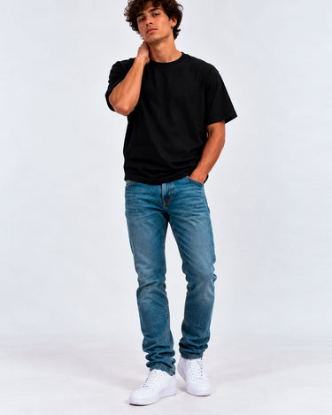 Pantalón vaquero azul claro en un hombre con camiseta negra y zapatillas blancas, fondo gris claro.