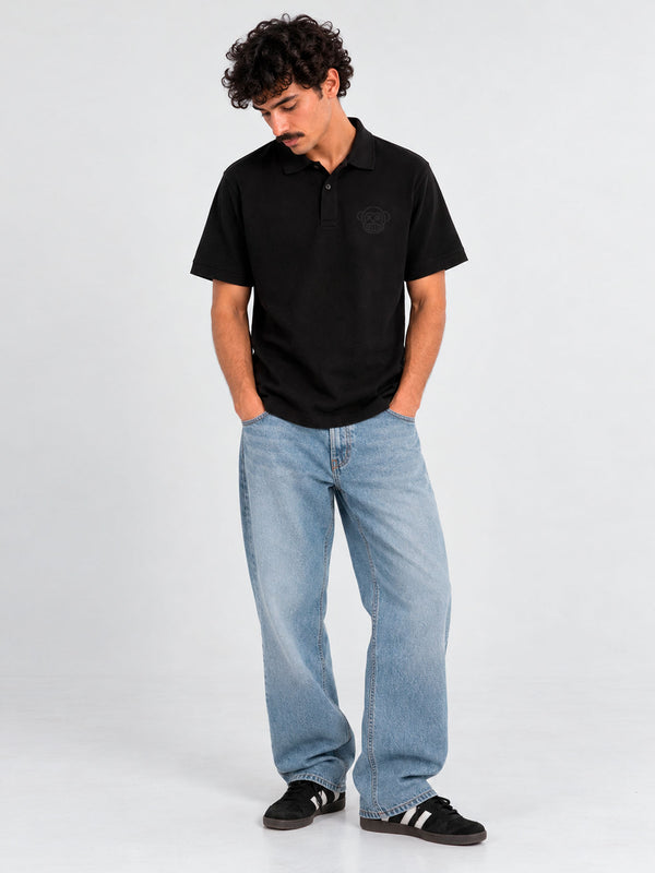 Polo negro de hombre con logo en el pecho, combinado con jeans claros y zapatillas negras.