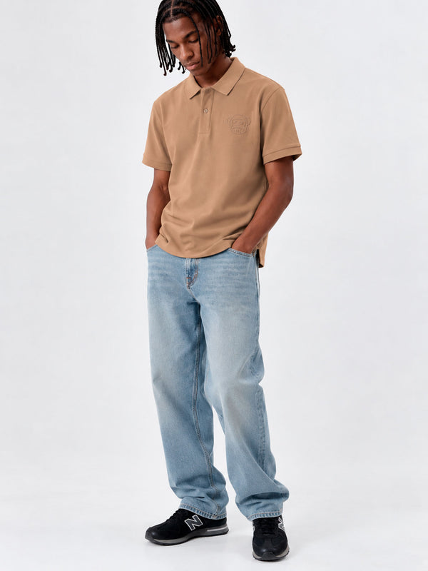Polo de hombre color caramelo con logo en el pecho, combinado con jeans claros y zapatillas negras.