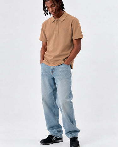Polo de hombre color caramelo con logo en el pecho, combinado con jeans claros y zapatillas negras.
