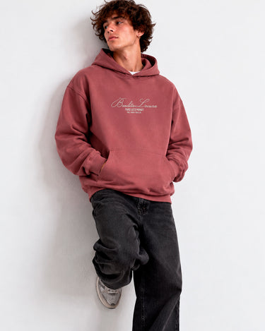 Sudadera unisex color vino con bordado blanco que dice 'Bendita Locura', modelo apoyado en la pared