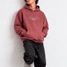 Sudadera unisex color vino con bordado blanco que dice 'Bendita Locura', modelo apoyado en la pared