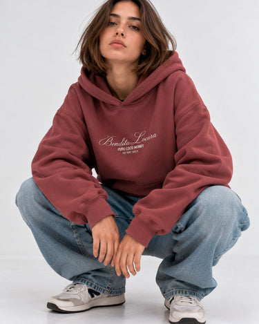Sudadera unisex color vino con bordado "Bendita Locura" y logo de Loco Monky, modelo sentado con jeans claros.