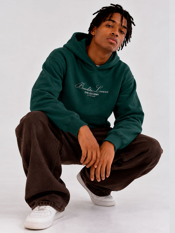 Sudadera unisex verde oscuro con bordado 'Bendita Locura' y logo de Loco Monky, modelo sentado con pantalones oscuros.