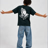 Camiseta negra con diseño de manos en la espalda, modelo de pie con jeans claros y brazos extendidos.