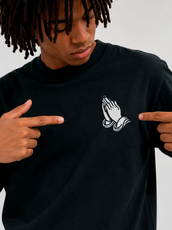 Camiseta negra con diseño de manos en oración en el pecho, modelo señalando el estampado.