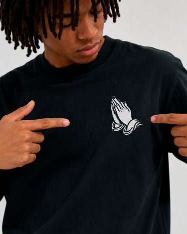 Camiseta negra con diseño de manos en oración en el pecho, modelo señalando el estampado.