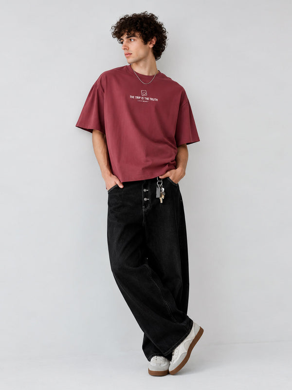 Camiseta boxy color púrpura con texto en el pecho, pantalones negros y zapatillas blancas, modelo de pie.