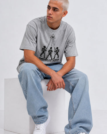 Camiseta gris con estampado de Godbusters, modelo sentado con pantalones anchos y zapatillas blancas.