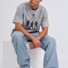 Camiseta gris con estampado de Godbusters, modelo sentado con pantalones anchos y zapatillas blancas.