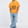 Camiseta amarilla con diseño de tres figuras y texto 'GODBUSTERS', pantalones jeans claros y zapatillas blancas.