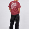 Camiseta de color burdeos con texto blanco en la espalda, hombre de pie con pantalones negros.