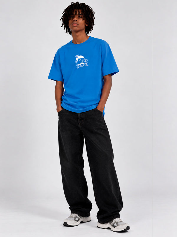Camiseta azul con diseño gráfico en el pecho, modelo de pie con pantalones negros y zapatillas deportivas.