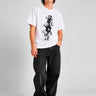 Camiseta blanca con diseño gráfico en negro, modelo masculino con pantalones oscuros y zapatillas blancas.
