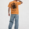 Camiseta mostaza con diseño de mono en la espalda, texto en negro, pantalones de mezclilla claros.