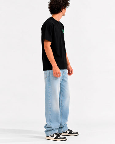 Camiseta negra con bordado en el pecho, modelo de hombre de pie, jeans claros y zapatillas negras.