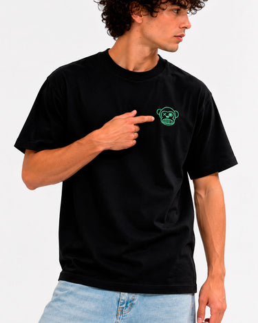 Camiseta negra con bordado verde de un mono en el pecho, modelo señalando el diseño.