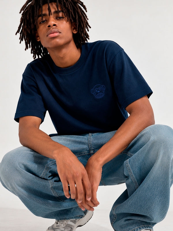 Camiseta azul oscuro con diseño de oso en el pecho, modelo sentado con pantalones claros y zapatillas deportivas.