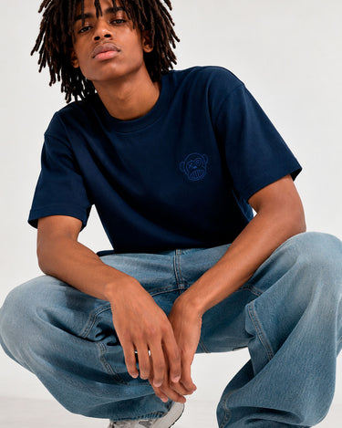 Camiseta azul oscuro con diseño de oso en el pecho, modelo sentado con pantalones claros y zapatillas deportivas.