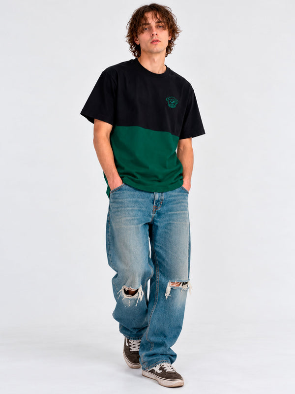 Camiseta de hombre en verde oscuro y negro, con logo en el pecho, combinada con jeans rotos y zapatillas.
