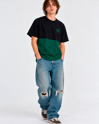 Camiseta de hombre en verde oscuro y negro, con logo en el pecho, combinada con jeans rotos y zapatillas.