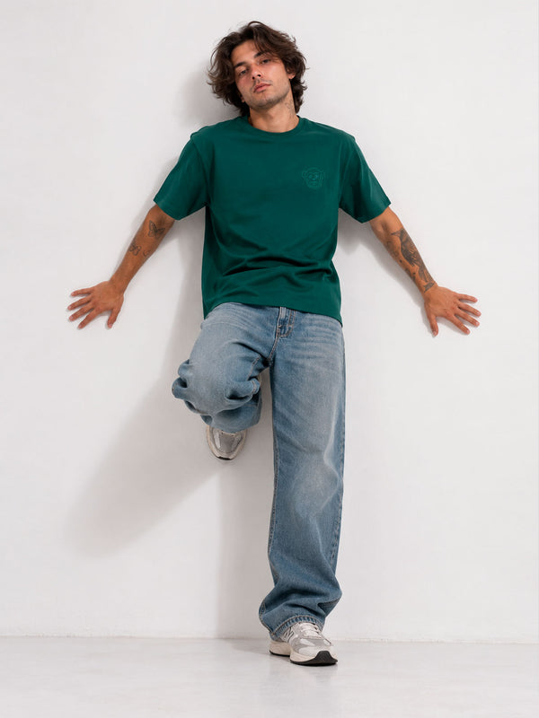 Camiseta verde oscuro con logo en el pecho, modelo con jeans claros y zapatillas, fondo blanco.