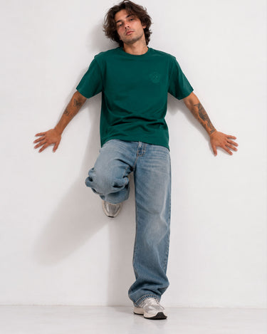 Camiseta verde oscuro con logo en el pecho, modelo con jeans claros y zapatillas, fondo blanco.