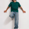 Camiseta verde oscuro con logo en el pecho, modelo con jeans claros y zapatillas, fondo blanco.
