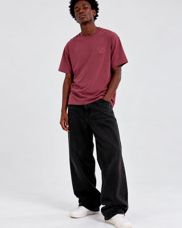 Camiseta de color púrpura con bordado en el pecho, modelo masculino con pantalones oscuros y zapatillas blancas.