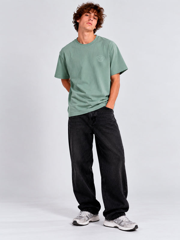 Camiseta verde con bordado en el pecho, combinada con pantalones negros y zapatillas deportivas blancas.