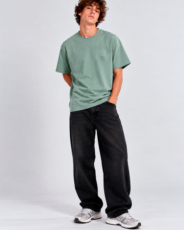Camiseta verde con bordado en el pecho, combinada con pantalones negros y zapatillas deportivas blancas.