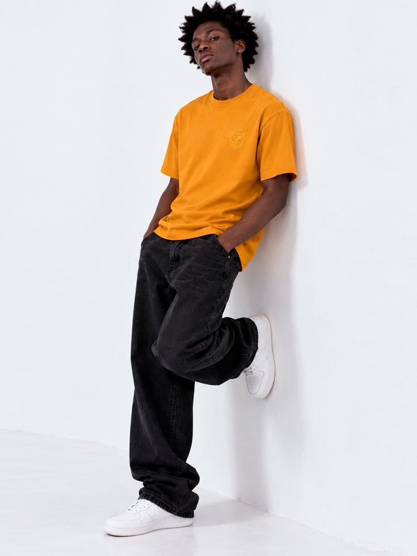 Camiseta amarilla con bordado en el pecho, hombre apoyado en la pared, pantalones negros y zapatillas blancas.