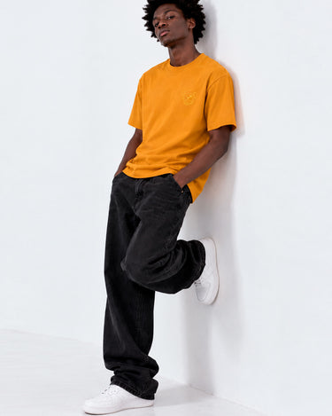 Camiseta amarilla con bordado en el pecho, hombre apoyado en la pared, pantalones negros y zapatillas blancas.