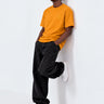 Camiseta amarilla con bordado en el pecho, hombre apoyado en la pared, pantalones negros y zapatillas blancas.