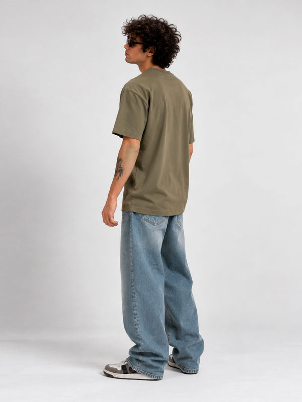 Camiseta de hombre color oliva, vista trasera, con pantalones anchos de mezclilla y zapatillas deportivas.