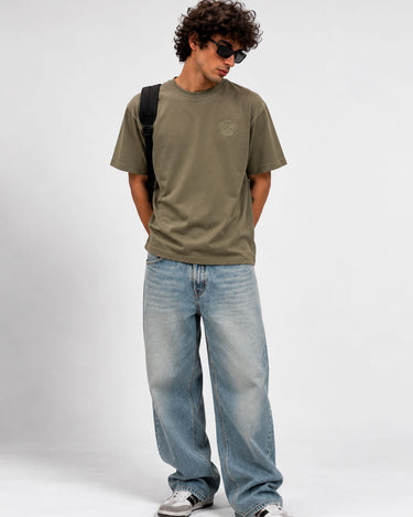 Camiseta verde oliva con logo en el pecho, modelo con pantalones claros y gafas de sol.