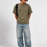 Camiseta verde oliva con logo en el pecho, modelo con pantalones claros y gafas de sol.