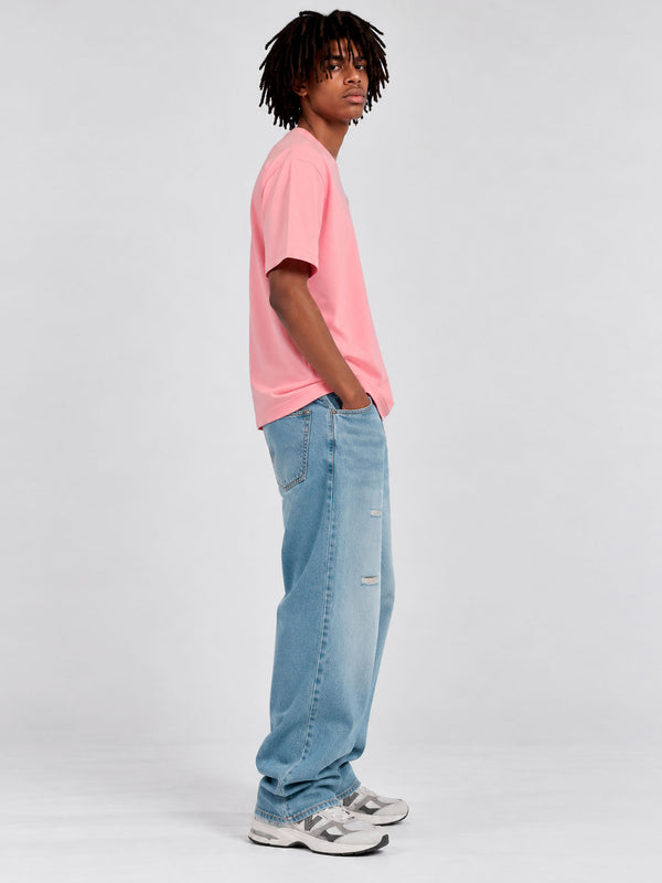 Camiseta rosa de hombre con pantalones de mezclilla claros, modelo de pie con cabello rizado.
