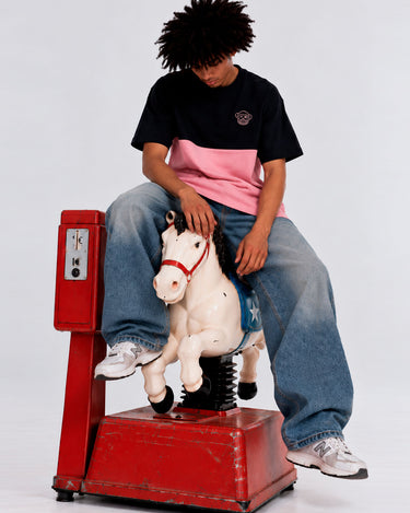 Camiseta de hombre en tonos rosa y negro, con logo en el pecho, modelo sentado en un caballito de madera.