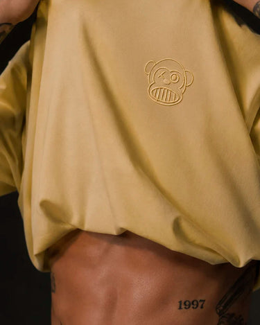 Camiseta amarilla suave con logo bordado de mono en el pecho, fondo oscuro y detalles de tatuajes visibles.