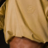 Camiseta amarilla suave con logo bordado de mono en el pecho, fondo oscuro y detalles de tatuajes visibles.
