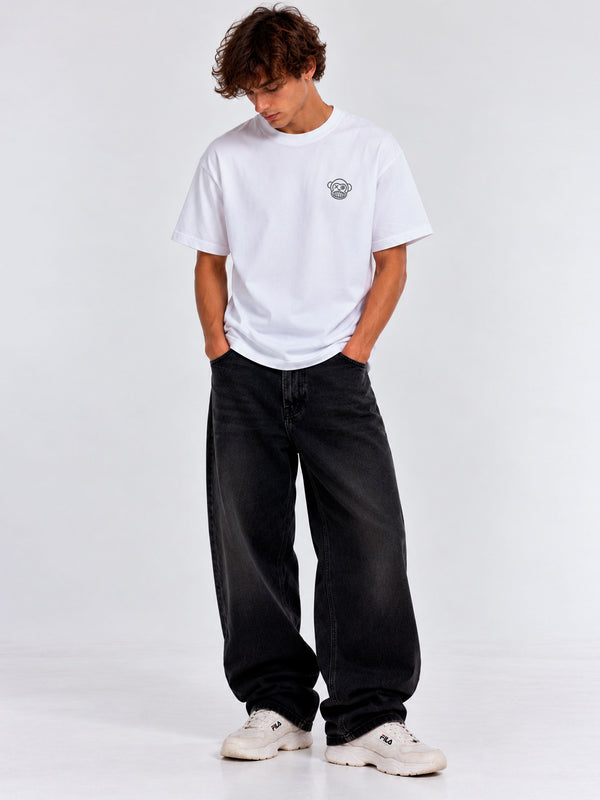 Camiseta blanca con logo en el pecho, pantalones oscuros y zapatillas blancas, modelo de pie con manos en los bolsillos.