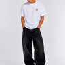 Camiseta blanca con logo en el pecho, pantalones oscuros y zapatillas blancas, modelo de pie con manos en los bolsillos.