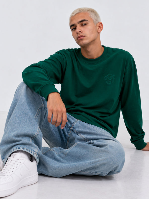 Camiseta de manga larga color verde oscuro, con logo en el pecho, combinada con jeans claros y zapatillas blancas.