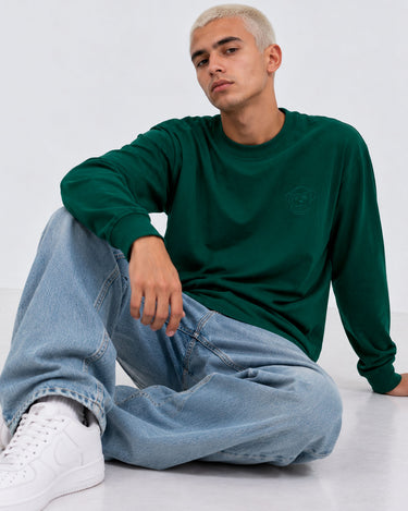 Camiseta de manga larga color verde oscuro, con logo en el pecho, combinada con jeans claros y zapatillas blancas.