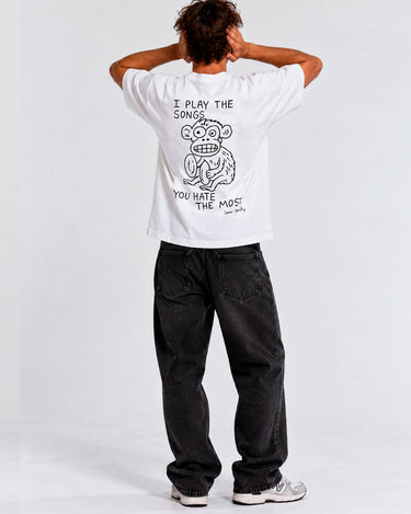 Camiseta blanca con diseño de mono y texto en la parte trasera, pantalones oscuros y fondo neutro.
