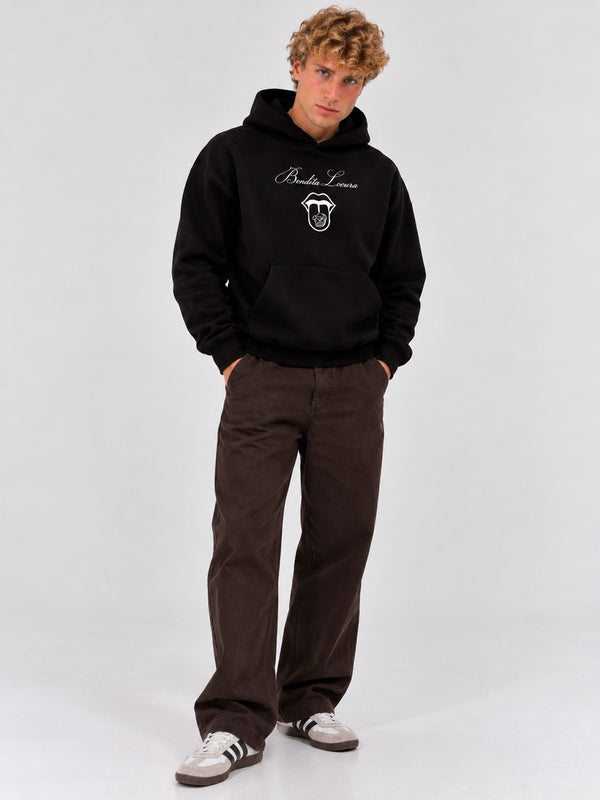 Sudadera negra con capucha y diseño en el pecho, pantalones marrones y zapatillas blancas con rayas.