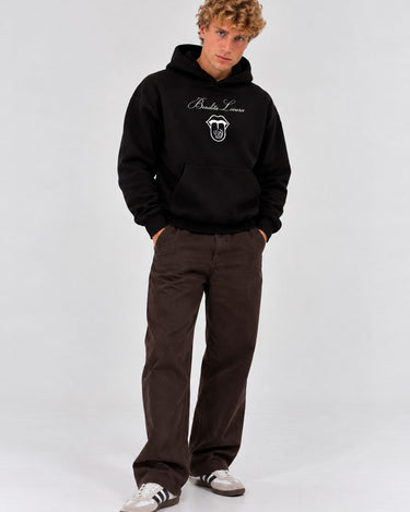 Sudadera negra con capucha y diseño en el pecho, pantalones marrones y zapatillas blancas con rayas.